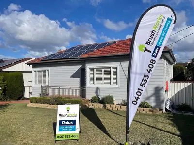 Single-storey house with grey weatherboards and white shutters, with a Brushworks flag and Dulux Accredited Painter sign on the front lawn
