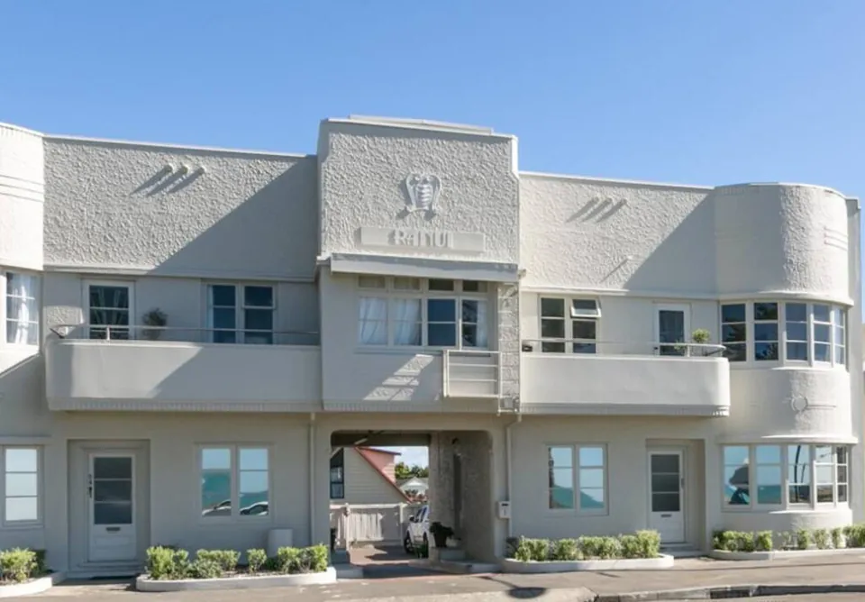 Two-storey art deco apartment building painted in soft grey with white trim, curved corner bays and a central archway