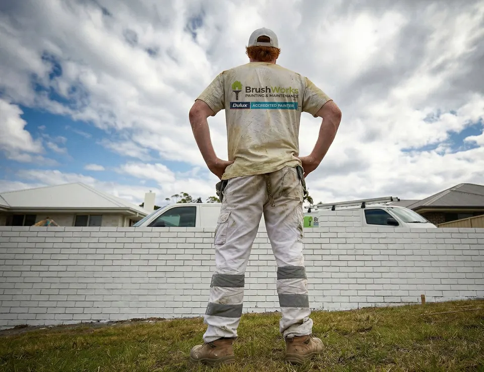Painter in white overalls with hands on hips looking at a freshly painted white brick fence under a cloudy sky