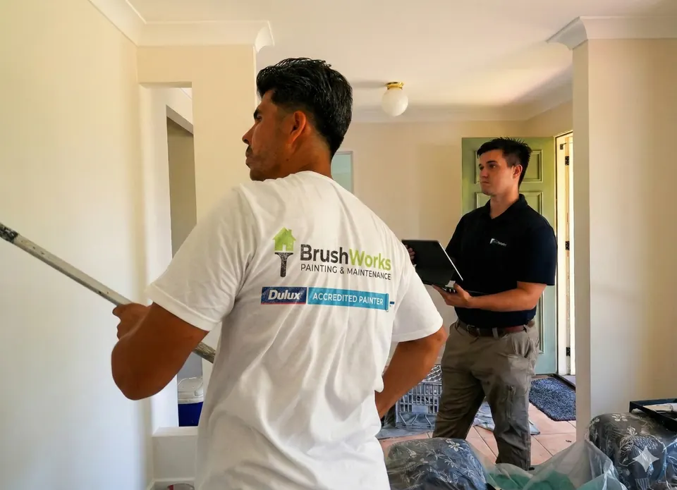 Brushworks painter holding an extension roller talks with a man holding a tablet inside a room being painted