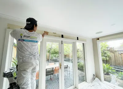 Painter on a step brushing the cornice above a row of white French doors in a bright room