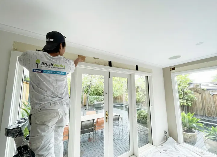 Painter on a step brushing the cornice above a row of white French doors in a bright room