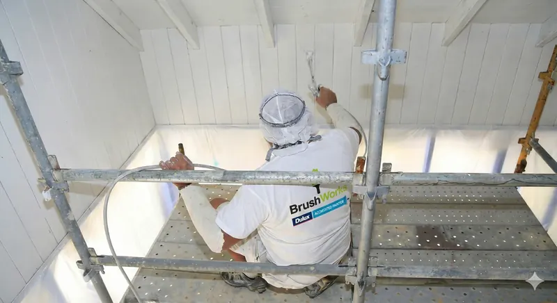 Painter in a spray hood working from scaffolding to spray a white timber-lined ceiling