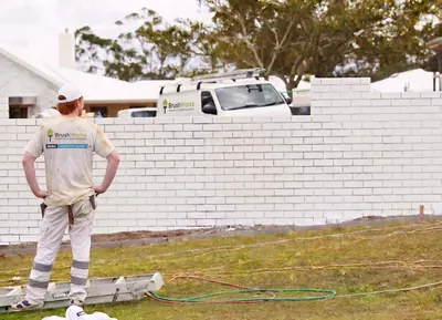Painter standing beside a freshly painted white rendered brick fence with a spray hose on the grass