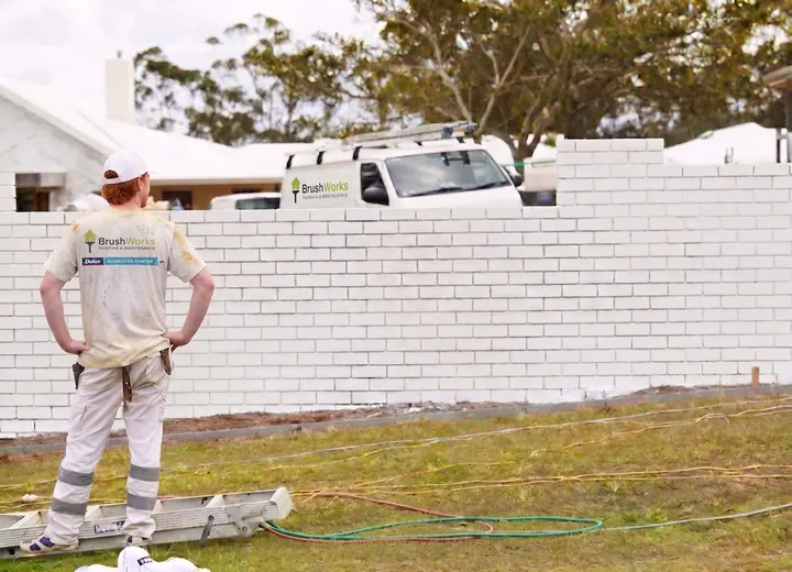 Painter standing beside a freshly painted white rendered brick fence with a spray hose on the grass