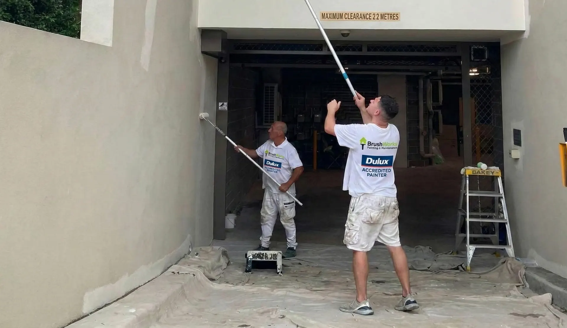 Two Brushworks painters using extension rollers on the walls and ceiling of an apartment building's vehicle entry, with drop sheets laid on the driveway