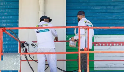 Two Brushworks painters on an orange mobile scaffold spray-painting blue and green weatherboard walls